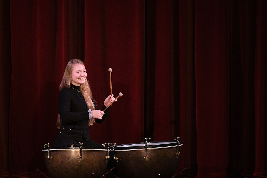 A female student playing the timpani drums, surrounded with by red curtains on stage.